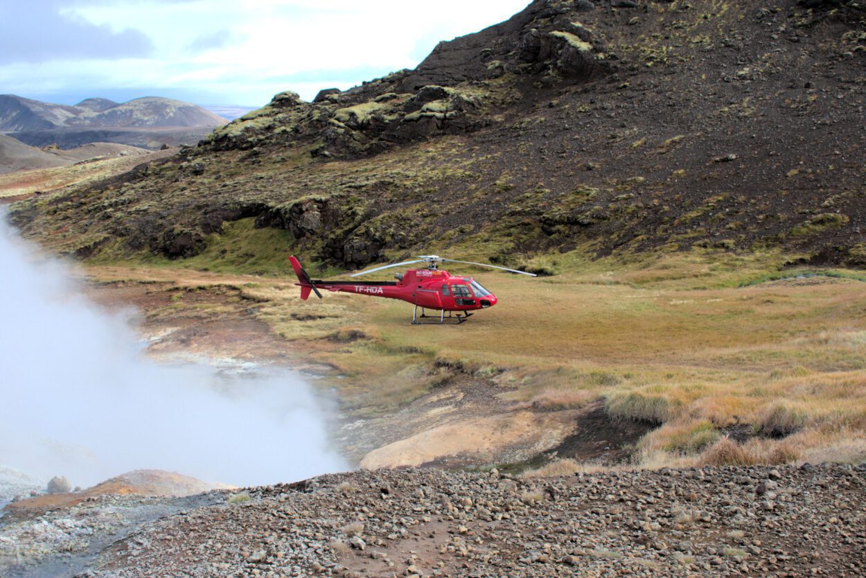 Ein roter Hubschrauber fliegt über einen Berg, aus dem Dampf austritt.