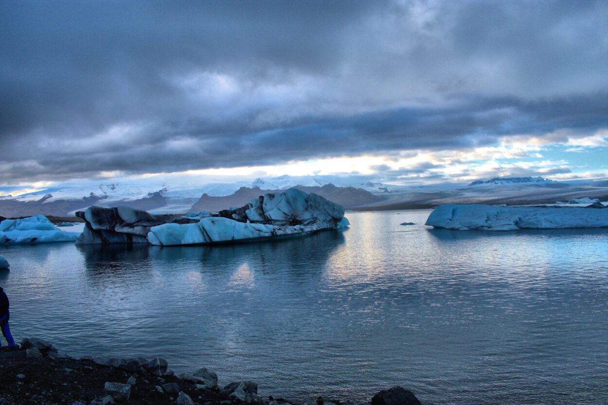 Eine Person steht in der Nähe eines Gewässers mit Eisbergen im Hintergrund.